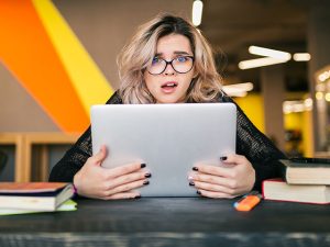 Retrato de uma jovem mulher bonita com expressão de pânico no rosto, sentada à mesa com um notebook demonstrado ter ansiedade de informação.