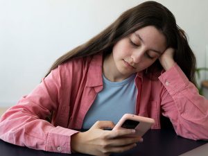 Garota em uma mesa, demonstrando expressão de tédio ao olhar o celular. A pressão social por meio das redes sociais desafia a autoaceitação.