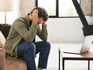 Homem sentado no sofá em frente ao notebook com as mãos na cabeça representando estar passando por uma crise de ansiedade.