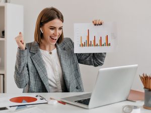 Mulher em frente a sua mesa de trabalho segurando um papel com um gráfico e comemorando ter conseguido lidar com a procrastinação e entregue o que foi planejado.