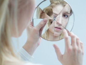 Mulher olha seu reflexo em um espelho trincado representando a ambiguidade entre sua imagem real e ideal.